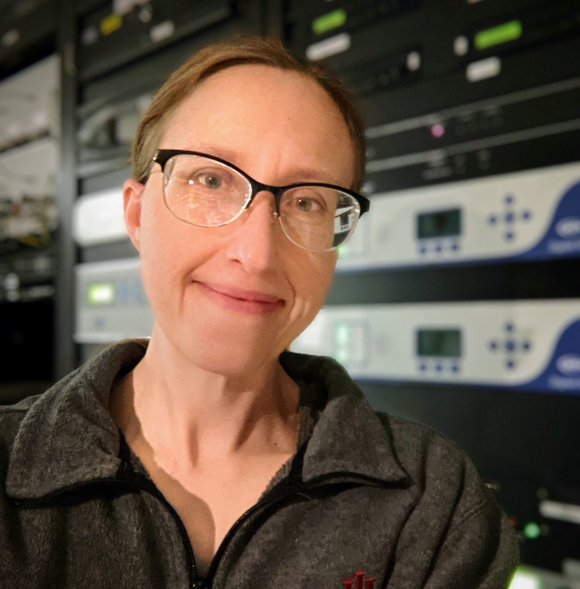 Headshot of a smiling woman (Kate Crum) standing in front of broadcast equipment.